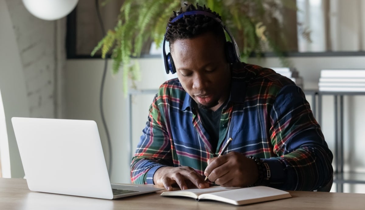 African man in a plaid shirt writing in a notebook in front of a silver laptop