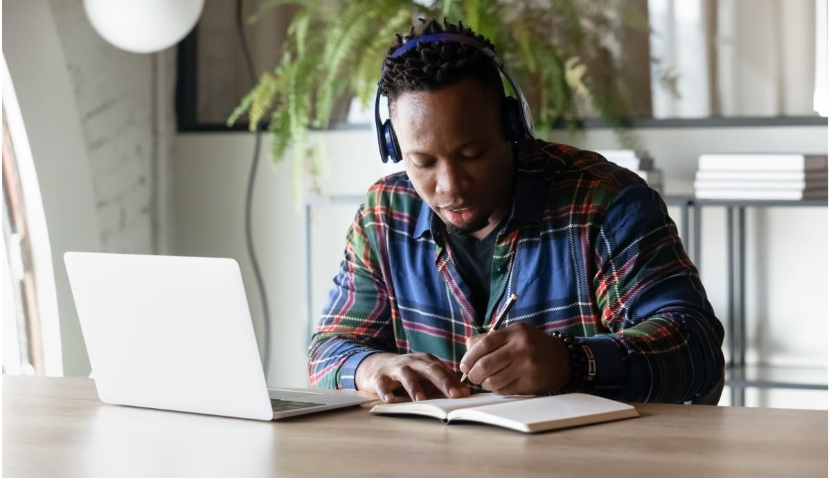 An African American man with short dreadlocks wears headphones and a plaid shirt while sitting behind a laptop computer. He appears focused and engaged in his work.